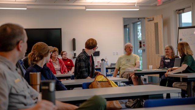 A group of people sitting look at a man speaking from his chair in the back of the room.