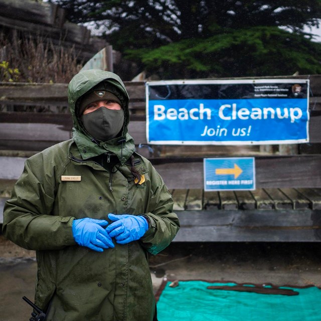 A park ranger in a hooded green coat stands in the rain in front of a blue banner. 