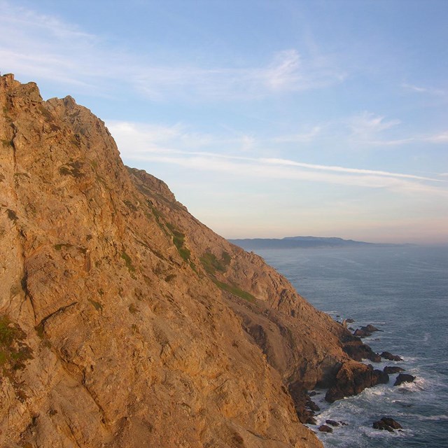 Golden hour light reflects off of a tall, rocky oceanic headland with wispy clouds in the sky.
