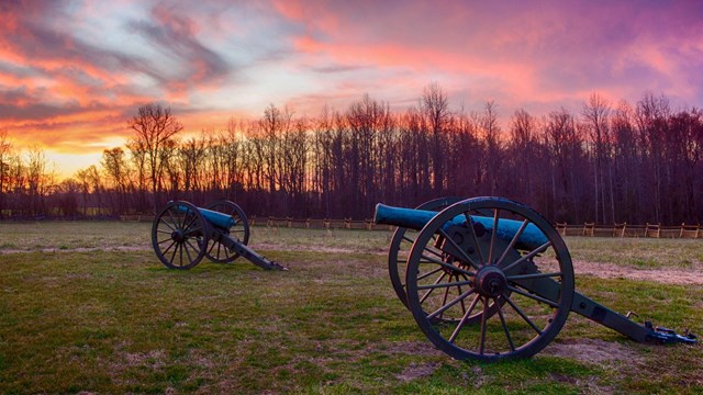 A pink and purple sunset behind two bronze Civil War cannons.