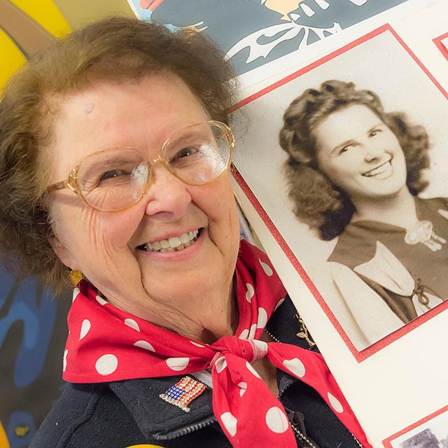 An older woman in an NPS volunteer uniform holds a self-made board with historic photos. 