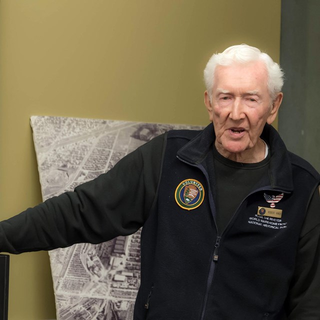 An older man is pointing at a self-made board with historic photos. 