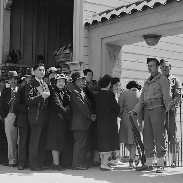 A historic photo of a group of Japanese Americans waiting in line. Soldiers stand in front.