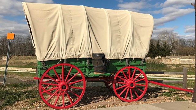 A covered wagon in a museum with a painted scene of a blue sky and grassy field behind it.