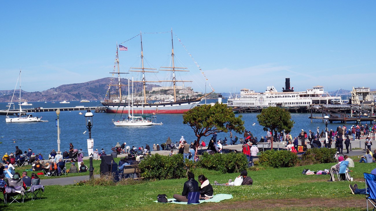 visitors sit on a grassy lawn with Aquatic Park Cove and Balclutha in the background