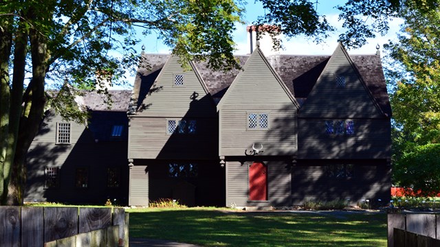 gray three-story house with red door and sun casting shadows of trees over 