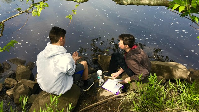 Two people sit at the edge of a river. 