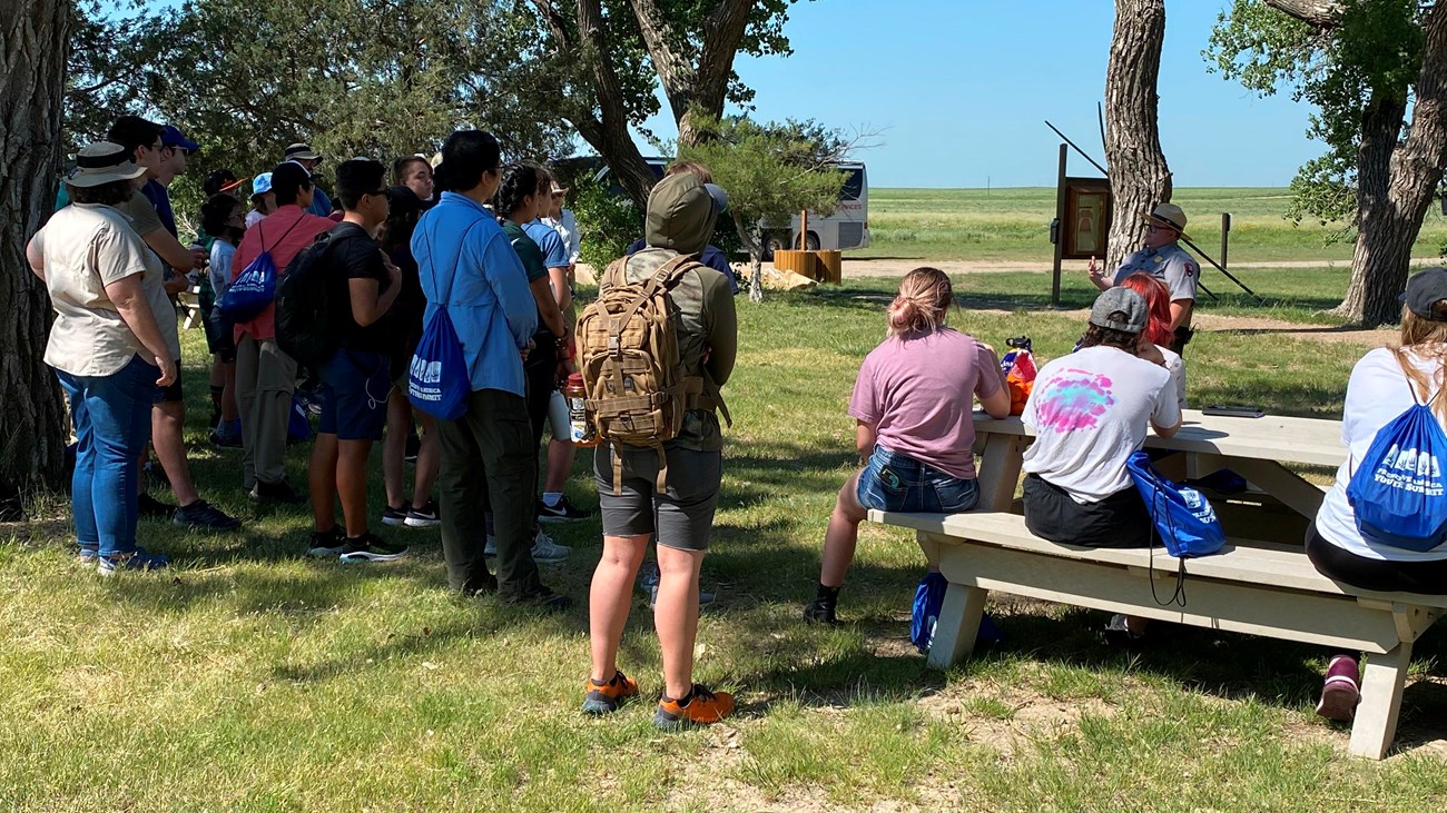 A park ranger speaks to youth standing in the shade of trees. 