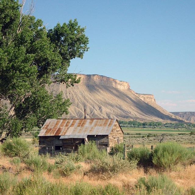 Historic building in a shrubland with large trees to the left and towering mesas in the distance.