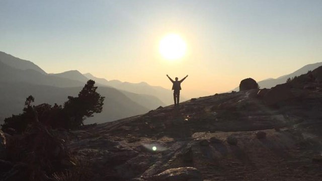 A person in silhouette stands on a rocky slope, reaching toward the sun just above them