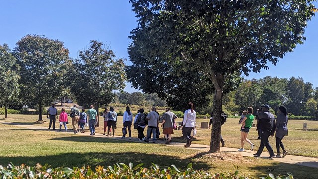 A crowd follows a tall ranger on a sidewalk framed by bright green grass.