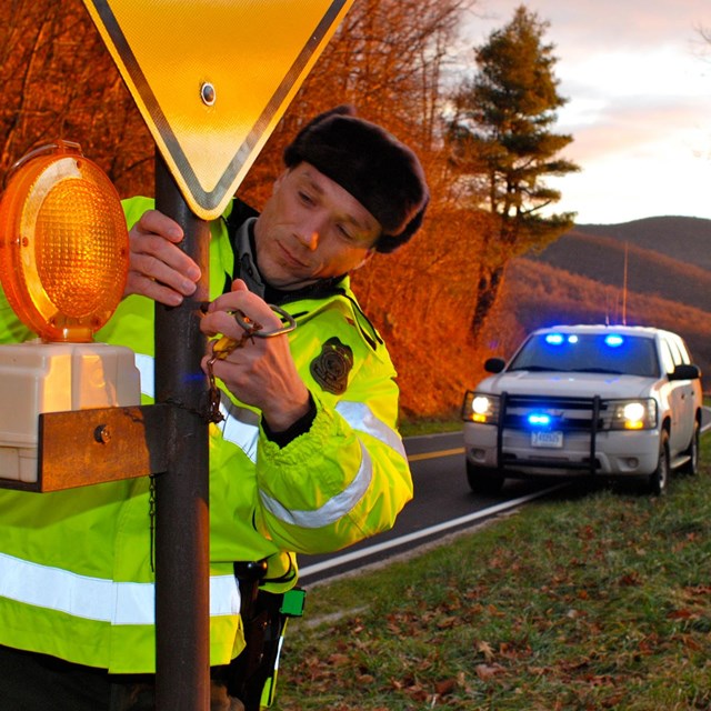 A law enforcement ranger closes a gate with the lights of his police car on in the background.