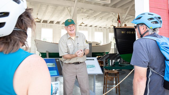 A man in a volunteer uniform stands inside a building and smiles at two people facing him.