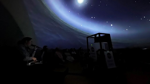 alt="A ranger uses a microphone to speak to a group of students under a projected starry night sky"