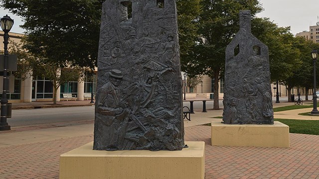 Two metallic appearing monuments that resemble burned structures stand on a brick walkway in a park.