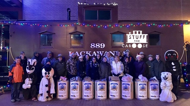 group of people and costume mascots with food drive buckets in front of holiday decorated caboose