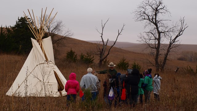 A small gathering of visitors gathers around a park ranger gesturing at a white prairie Teepee