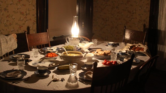 A lamplit table with 6 chairs appears to have food and be laid out for a meal.