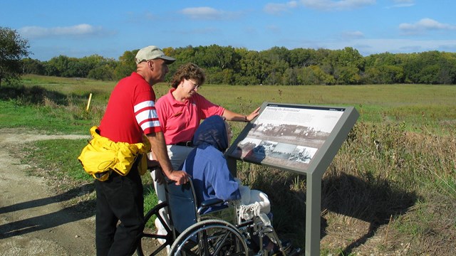 3 park visitors look at an informational wayside exhibit. One of the visitors is in a wheelchair.