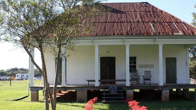 White cabin with red roof behind two rows of red flowers on a green lawn.