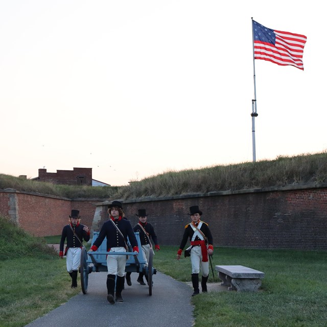 Living historians walking out of Fort McHenry, pulling a wagon, with the 17x25 flag flying