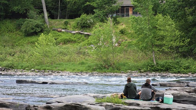 Couple sits on rocks on the edge of a river. Yellow-green trees form a forest across the river.