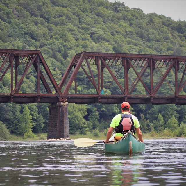 Man in life jacket sitting in canoe on the river with a bridge and forested hills in background.