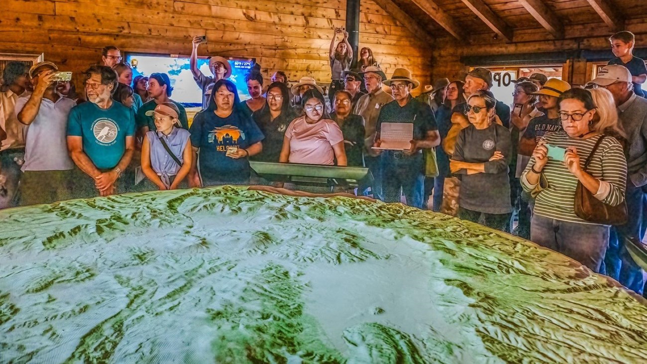 A crowd gathers around a large, circular, 3D map of a caldera.