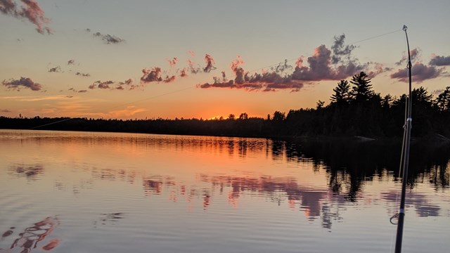 A fishing pole casts a line from a boat on a scenic lake at dusk.