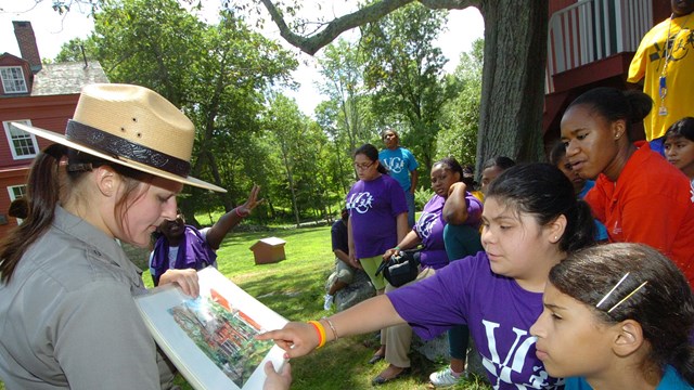 A Park Ranger holds an image of a painting at Weir Farm and students point at it in excitement