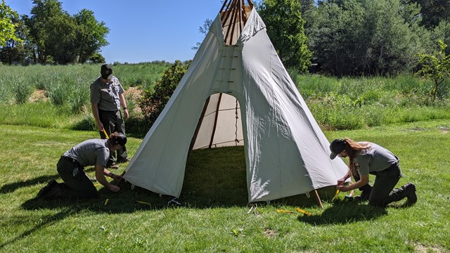 Three rangers gathered around a tipi putting stakes around its base