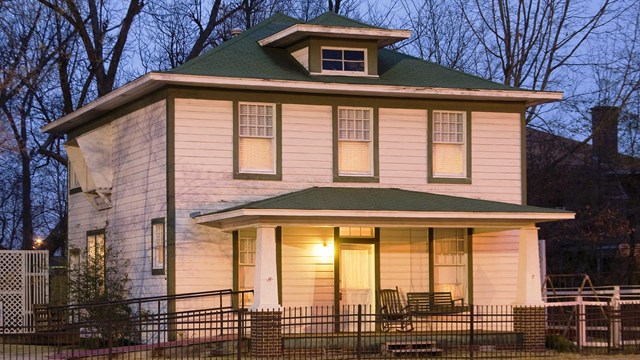 White two-story house with green trim lit at dusk with trees behind