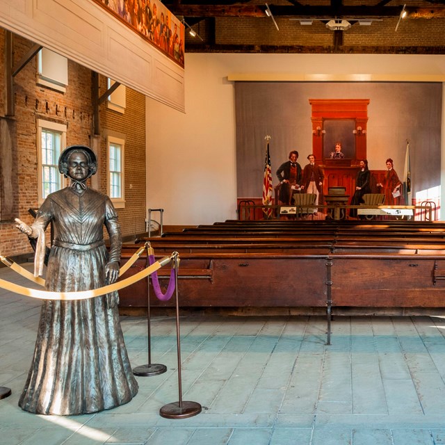 An empty room full of benches, with a statue of a woman welcoming in the foreground.