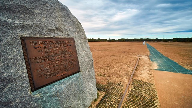 A picture of a large boulder with a bronze plaque on it and a pathway leading to 4 other markers
