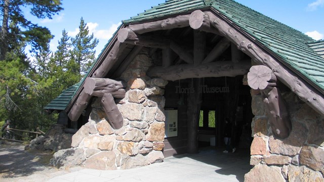 A wood and stone building with a sloped roof sits amongst conifer trees.