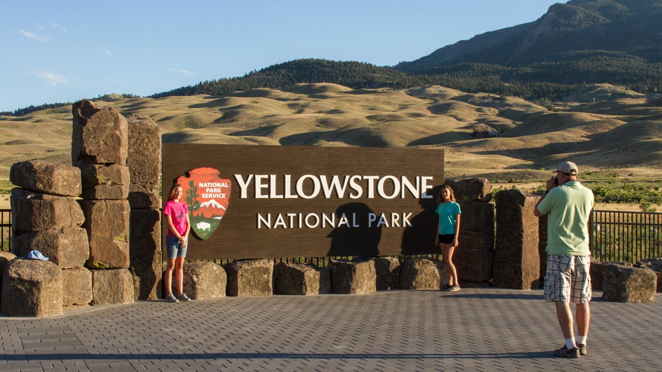 Two girls stand in front of a large Yellowstone National Park sign while their dad photographs them