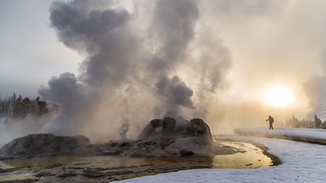 A skier enjoys a winter sunrise at near a steaming geyser.