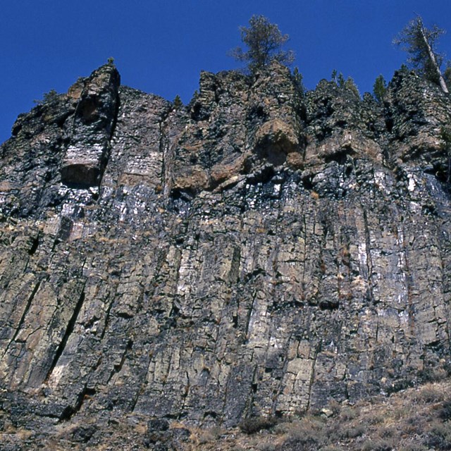Brown and gray columns of rock make up a cliff that towers up to a deep blue sky.