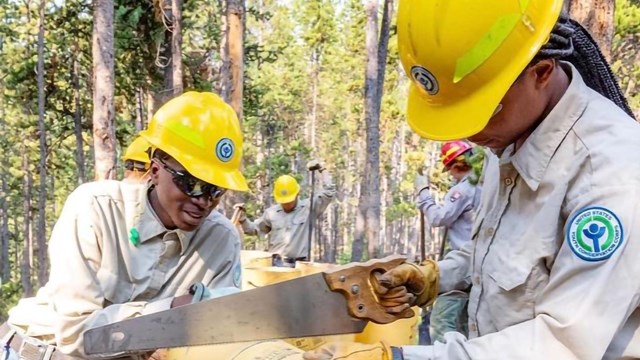 Two youth in yellow hard hats use a saw to cut a tubes