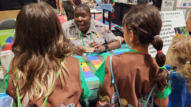 Two youth wearing brown vests with Girl Scout patches stand in front of a table with a Park Ranger.