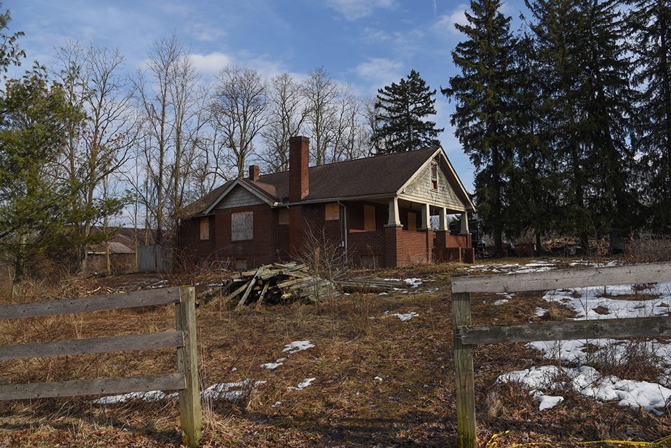 Beyond a fence is a boarded up brick house with evergreen trees, right, and barn, left.