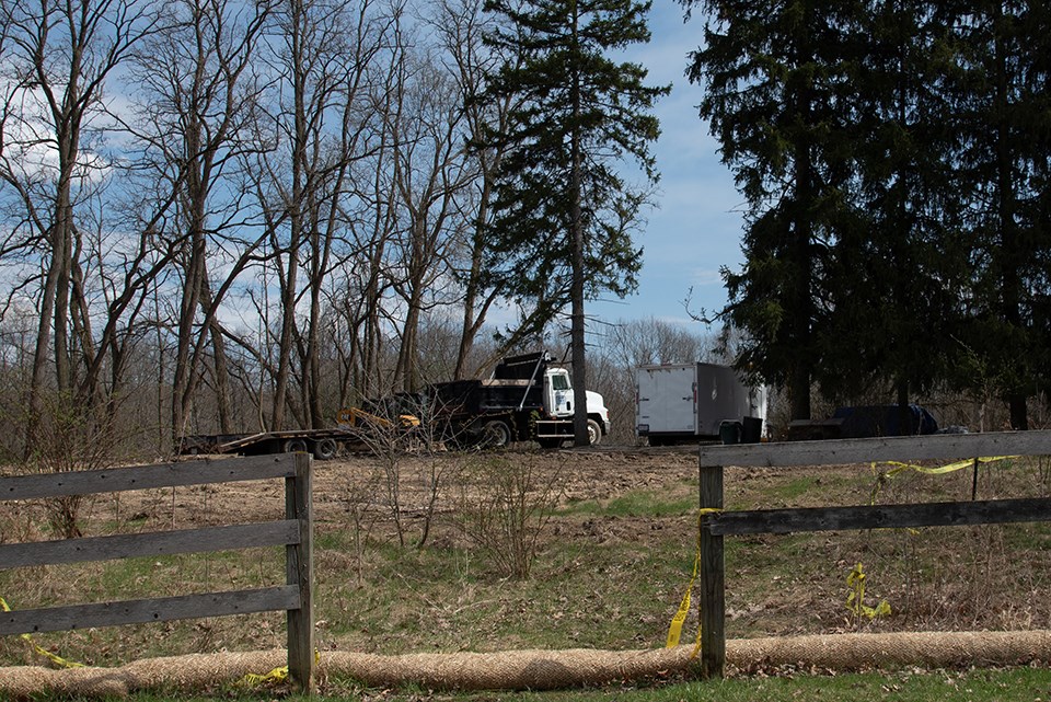 Beyond a fence is a boarded up brick house with evergreen trees, right, and barn, left.