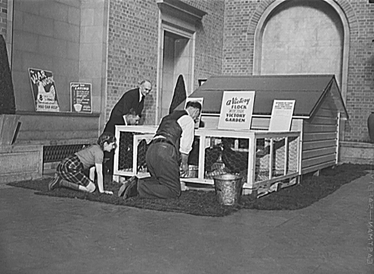 Black and white photo of a white man in a tie kneeling in a garden with plants. Three white girls in Girl Scout uniforms look on.