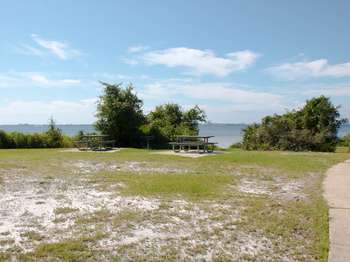 Two picnic tables on green grass in the foreground. Green trees, blue water, and blue sky in the background.