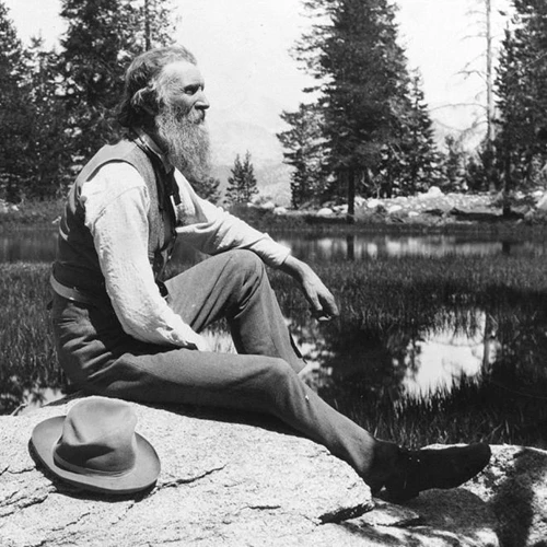John Muir sits on a rock by a pond or lake. His hat lies on the rock next to him.