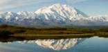 pond surrounded by green brush, reflecting a distant range of snow-covered mountains that are dominated by one massive mountain