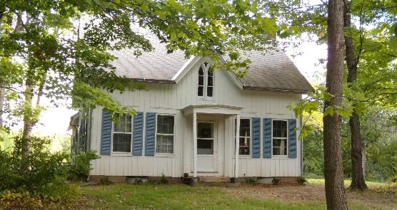 Welton farmhouse, a small, white, and modest single story home with a pointed roof and blue shutters, sits among green trees.