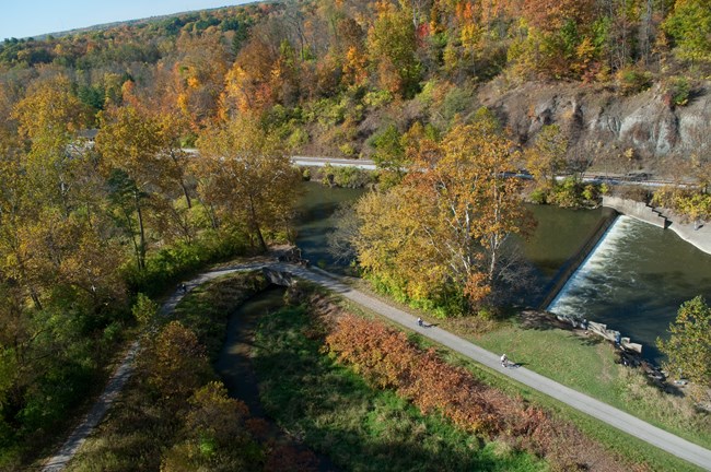 A lowhead dam on a green river viewed from above, surrounded by orange, yellow and green fall tree colors.