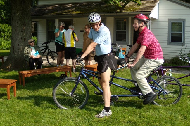 Two men wearing helmets, one standing and one seated on a tandem bike, and other cyclists in front of a white house.