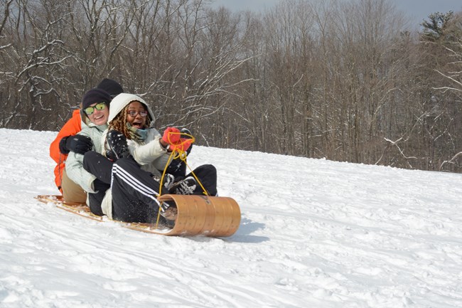 Three people in winter coats and gloves smile as they ride down a snowy hill on a wooden toboggan.